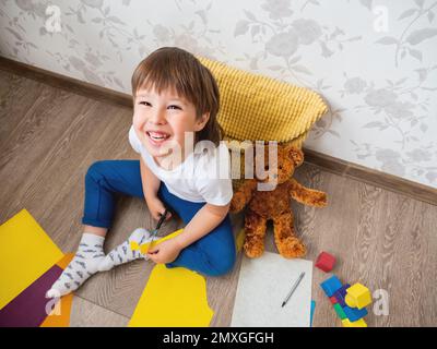 Toddler boy learns to cut colored paper with scissors. Kid sits on floor in kids room with toy blocks and teddy bear. Educational classes for children Stock Photo