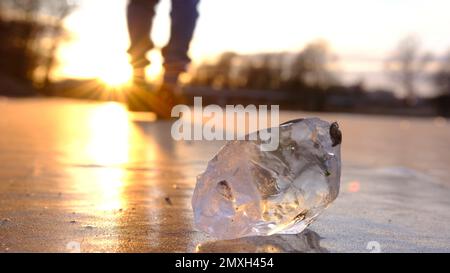 The lonely figure of a blured man walking on the frozen river at sunset, with peace of crystal ice in forward line, the reflection of the sun. Stock Photo