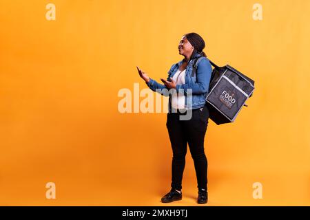 Deliverywoman with takeout thermal backpack showing takeaway advertisement in studio with yellow background. Restaurant employee delivering fast food order to clients during lunch time Stock Photo