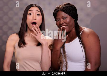 Two diverse surprised women laughing with hand over mouth, hearing funny joke. African american and asian beautiful girls giggling with shocked facial expression portrait Stock Photo