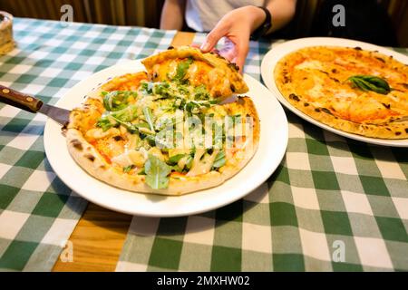 Female hands taking slices of authentic Italian caesar stone oven pizza with arugula, sauce, grilled chicken, herbs and spicy parmesan cheese. Stock Photo