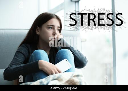 Word STRESS and depressed young girl sitting at window indoors Stock Photo