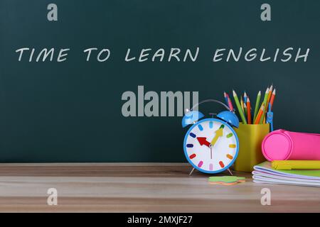Alarm clock and stationery on table near green chalkboard with text Time to Learn English Stock Photo