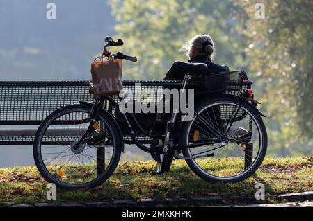 A lady enjoys the warming autumn sun on a park bench in the Olympic Park Munich, Bavaria, Germany, Europe Stock Photo