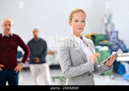 Managing the factory with poise and confidence. A manager using a tablet during a factory inspection with her coworkers in the background. Stock Photo