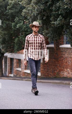 Urban cowboy. a handsome man wearing a check shirt and cowboy hat walking along a road. Stock Photo