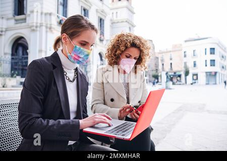 Two businesswoman with face masks using mobile phone and laptop while sitting outdoors. Stock Photo