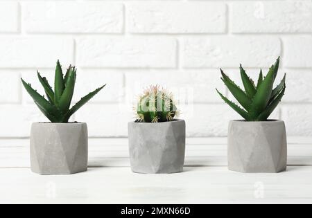 Beautiful artificial plants in flower pots on white wooden table near brick wall Stock Photo