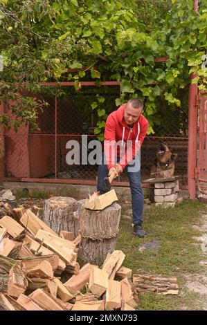 A young man in the village cuts firewood with an axe to heat his house in winter. Stock Photo