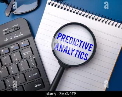 Wireless keyboard and magnifying glass with the word PREDICTIVE ANALYTICS on blue background. Stock Photo