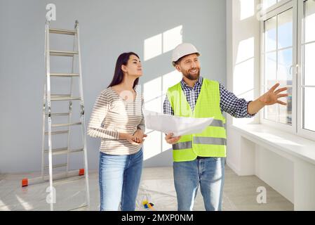 Smiling builder in uniform holding construction plan and talking to young woman house owner Stock Photo