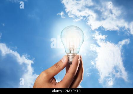 Solar energy concept. Man holding glowing light bulb against blue sky with clouds, closeup Stock Photo
