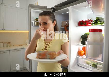 Young woman with spoiled sausage near open refrigerator in kitchen Stock Photo