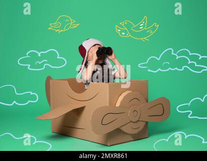 Cute little child playing in cardboard airplane on green background with illustrations Stock Photo