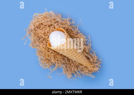 Waffle ice cream cone with a hidden white Easter egg in a paper nest on a blue background, concept, minimalism. Creative Easter Card Stock Photo