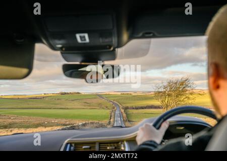 A man driving in the car on the empty countryside road alongside the green fields during the daytime Stock Photo