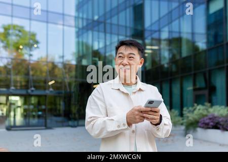 Smiling asian man in casual shirt walking in city using app on smartphone, man looking to side on background of office building from outside. Stock Photo