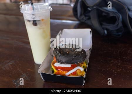 Burgers and Beverages: A Burger with Black Bun on a Wooden Table Next to a Drink Stock Photo