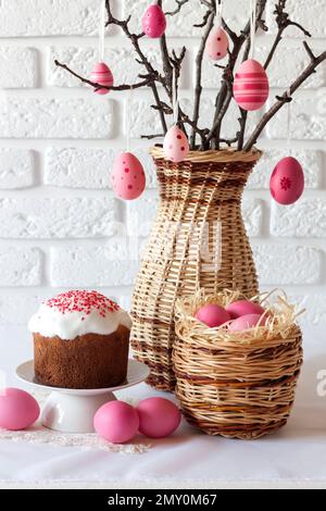 Easter composition with decorated tree branches in a wicker vase, pink colored eggs in wicker basket and Easter cake on white background Stock Photo