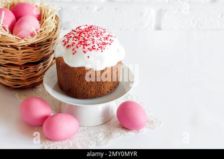 Minimalistic Easter composition with wicker basket with pink colored eggs and Easter cake on white background. Copy space Stock Photo