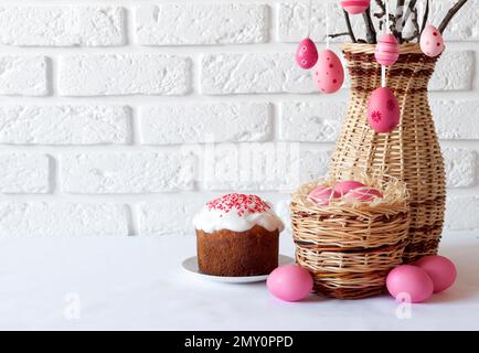 Easter composition with decorated tree branches in a wicker vase, pink colored eggs in wicker basket and Easter cake on white background. Copy space Stock Photo
