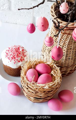 Easter composition with decorated tree branches in a wicker vase, pink colored eggs in wicker basket and Easter cake on white background Stock Photo