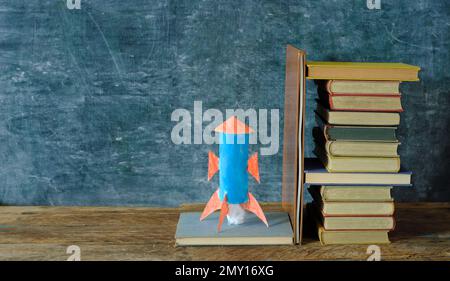 Stack of hardback books with rocketship launching on blackboard ...