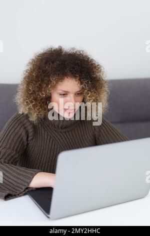 Beautiful curly woman working on notebook computer at home. Pretty young adult female person typing text on modern laptop Stock Photo