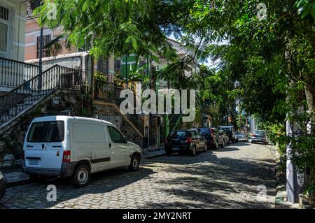 Cars parked on Ladeira de Santa Teresa cobblestone slope street in front of some houses in Santa Teresa district under summer afternoon sunny blue sky Stock Photo