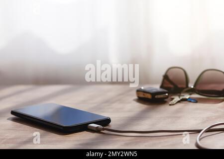 Mobile phone charging over a table with usb with cable. On background, defocused, there are car and home keys and sunglasses. Stock Photo