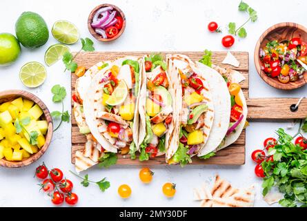 Tacos with grilled chicken, tomato salsa sauce, mango, cilantro and onion in tortilla on cutting board. White table background, top view Stock Photo