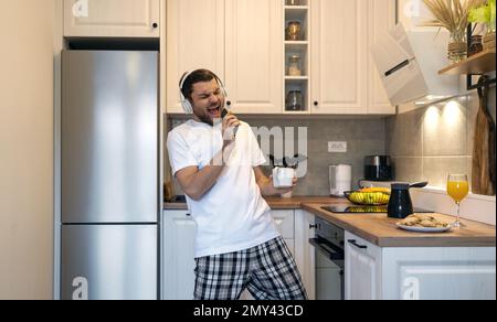 Young man in headphones singing in kitchen in morning using his mobile phone as a microphone. Stock Photo