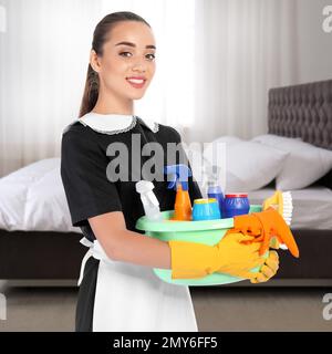 Beautiful chambermaid with cleaning supplies near bed in hotel room Stock Photo