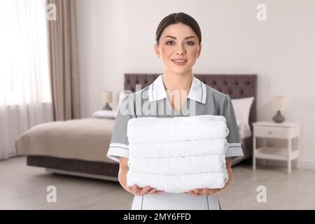 Beautiful chambermaid with clean folded towels near bed in hotel room Stock Photo