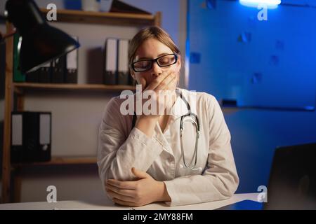 Tired woman doctor works on a laptop computer in the clinic. Female doctor in a white coat yawns and wants to sleep working at night. Stock Photo