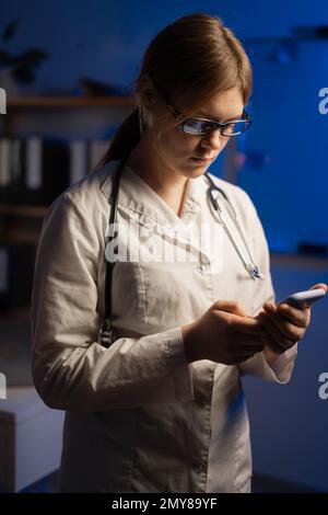 Shot of pretty female doctor using her mobile phone while standing in medical consultation during night shift. Stock Photo