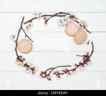 Decorative frame of flowers and macaroons on a white wooden background. Almond biscuits and branches of flowering apricots. Template for design. Flat Stock Photo