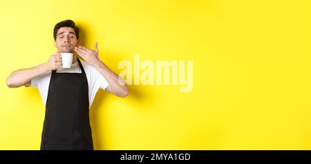Barista enjoying smell of coffee in mug, standing pleased with eyes closed, wearing black apron Stock Photo