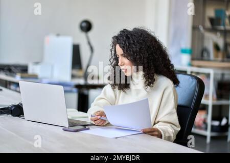 Young latin business woman holding documents working with laptop. Stock Photo