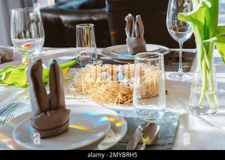 Beautiful Easter table setting with festive decor indoors. Napkin in shape of rabbit ears, colorful eggs in nest, fresh yellow tulips in vase. Sunligh Stock Photo
