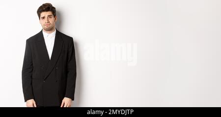 Image of gloomy and sad handsome male model in black suit, frowning and looking tired at camera, standing against white background Stock Photo