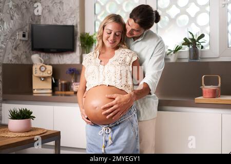 Loving caucasian couple at home in kitchen feeling baby kick together. Pregnant mom and Smiling dad Stock Photo