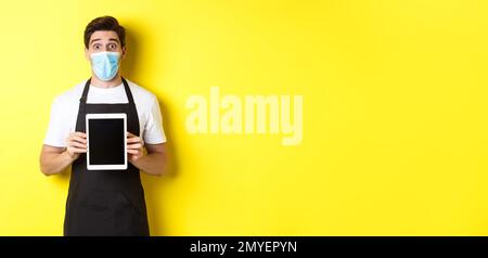 Concept of covid-19, small business and pandemic. Surprised waiter in black apron and medical mask showing tablet screen, standing over yellow Stock Photo
