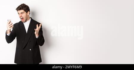 Portrait of angry businessman in black suit yelling at mobile phone, having an argument on video call, standing mad over white background Stock Photo