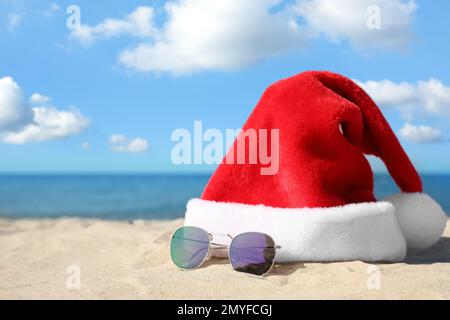 Santa's hat and sunglasses on sandy beach, space for text. Christmas vacation Stock Photo