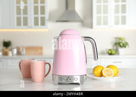 Modern electric kettle, cups and lemons on table in kitchen Stock Photo