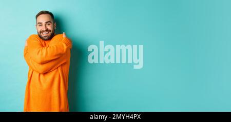 Happy man feeling comfortable, wearing warm sweater and hugging himself, smiling satisfied, standing over light blue background Stock Photo