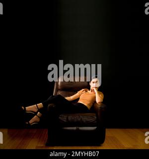 Beautiful Young Asian Woman Reclining on a Brown Leather Chair Against Black Backdrop Stock Photo