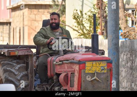 A smiling man, cell phone in hand, guides his old, red tractor through the streets of a village in northern Iraq. Stock Photo