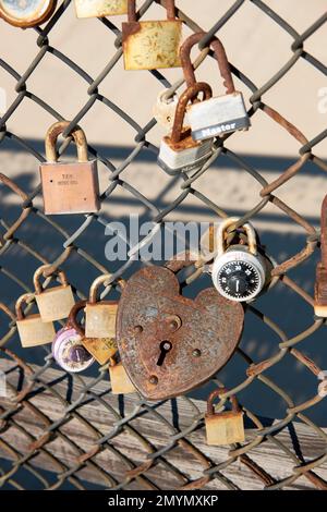 A rusty, heart shaped love lock padlock hangs on a chain link fence. Stock Photo